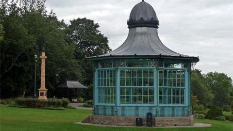 There is a bandstand in the field of a park which has green iron work and a dome roof. In the background is a tall column made from sandstone
