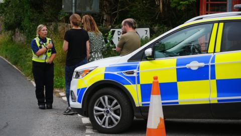 A female police officer in uniform talks to local residents standing next to a police car in Wheddon Cross in Somerset after a coach crash