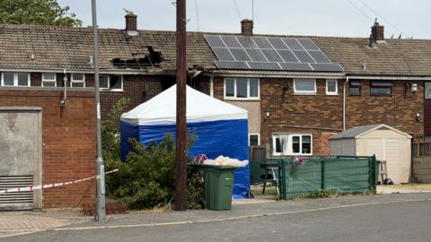 A blue and white police tent at the scene. A hole has been burnt into the roof of a terrace house.