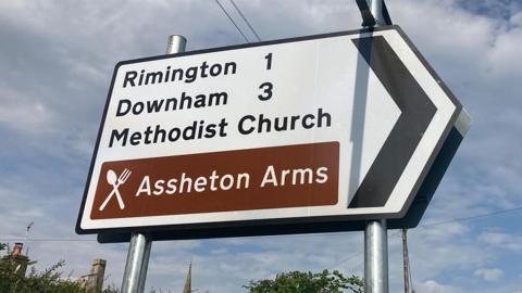 A white street sign with black text, pointing to the right saying "Rimington 1, Downham 3, Methodist Church" and a brown box of text saying Assheton Arms against a blue cloudy sky