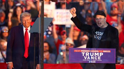 Trump and Musk on stage behind bulletproof glass 