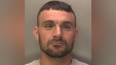 A police mugshot shows a man with brown hair and stubble. He is wearing a grey t-shirt.