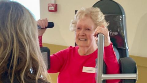A woman with back to us with the words 'Health Practitioner' on her sweat shirt and she is standing in front of an older woman who is sitting on a piece of rehabilitation equipment used to exercise her arms. 