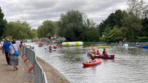 The River Great Ouse in Bedford town centre, with the promenade on the left. There are barriers up as it is the summer River Festival. Pedestrians walk along the promenade, and many canoes and kayaks are on the river. Children and adults wearing helmets paddle around, and there are weeping willows and other trees along the banks.