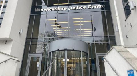 A pic of the outside of a court building. It is glass fronted with Warwickshire Justice Centre written in grey lettering high above a revolving door.
