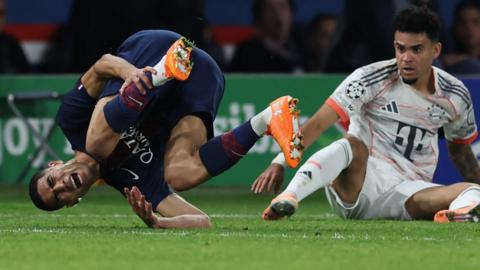 Achraf Hakimi in dark blue PSG kit clutches his left shin and closes his eyes as he screams in pain following a challenge from Bayern Munich's Luis Diaz during a Uefa Champions League match. Hakimi is almost curled up in a ball as he tumbles across the green turf, with Diaz sitting on the ground behind watching following the red card challenge