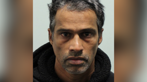 Custody image of Vas. He has short dark grey hair, grey stubble, and dark shadows under his eyes. 