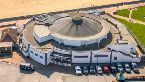 A drone image of the Ocean Room, showing the rotunda and it's roof. Next to it are the beach in the top of the image, and lawns and the bandstand to the right.