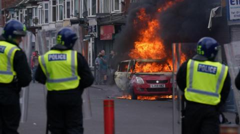 The backs of three police officers are visible as they stand a safe distance from a burning red car on a street. A row of shops can be seen in the distance. A group of people stand beyond the blazing car.