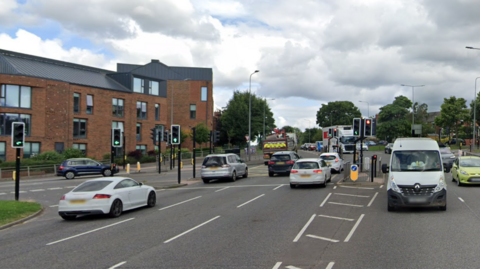 Neville's Cross in Durham is busy with vehicles at the junction on the A167. The traffic lights are on green and vehicles are heading through them. A three-storey brick building - which could be flats or offices such is the relative blandness of the design - with a zinc or copper-style pointed roof is to the left of the junction.