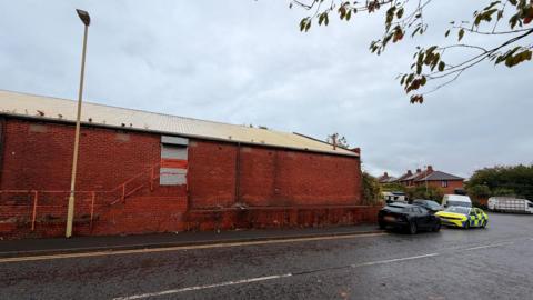 Photo of red brick industrial site with black car and police car in right hand corner of the photo on the street. The red brick building has a door with grey shutters and there are houses in the background. It is a grey day. 