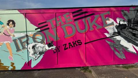 A pink hoarding with silver lettering depicting The Iron Duke by Zaks. Images bleeding from elsewhere in the hoarding are visible on the extremes of this picture.