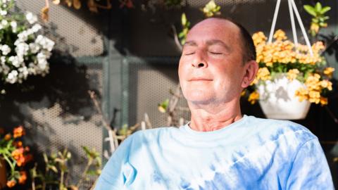 A man in a light blue t-shirt sitting outside with his eyes closed. The sun is on his face and there are hanging baskets of flowers in the background.
