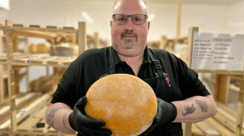 Steve Reid looks directly at the camera holding up a large cheese. He is wearing a black polo shirt, black apron and gloves. He has a number of tattoos on his arms and he is wearing glasses and has a moustache. He is standing in front of some wooden shelving. 