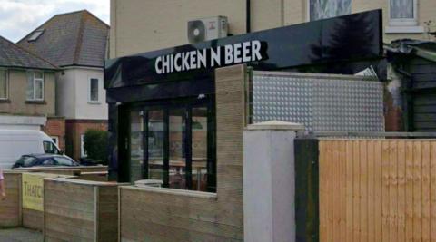 Google Street View of Chicken n Beer cafe. A black and glass fronted cafe on a street corner with an outside seating area separated from the pavement by wooden barriers.