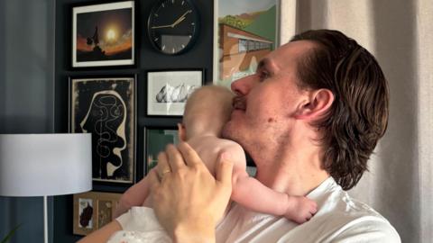A man with brown hair and a moustache holds a baby to his shoulder