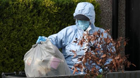 A forensic officer in a blue suit carries a bin bag in front of a hedge