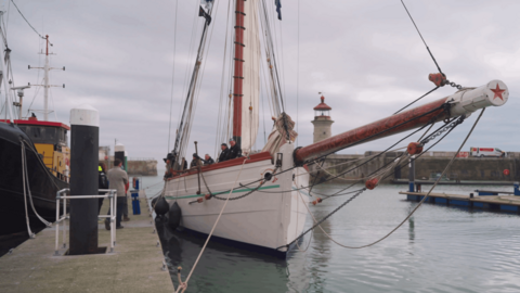 A white tall ship moored to a quay at Ramsgate harbour. Its sails are down and there are some people stood on the deck and on the quayside.