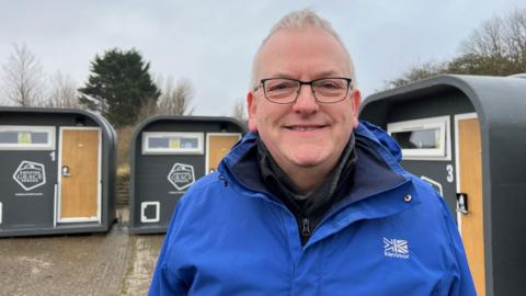 A man smiles has he stands in front of three small shed-like sleeping pods. He has grey hair and is wearing a blue coat and glasses. The pods are dark grey with light-brown wood-effect doors, a white-framed window and a logo reading Amazing Grace Spaces. 