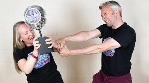 Kelly and Rob and standing next to each other wearing black t-shirts that say "dance floor heroes 2025". Kelly is holding onto a trophy shaped like a disco ball. Rob is trying to wrestle it off her