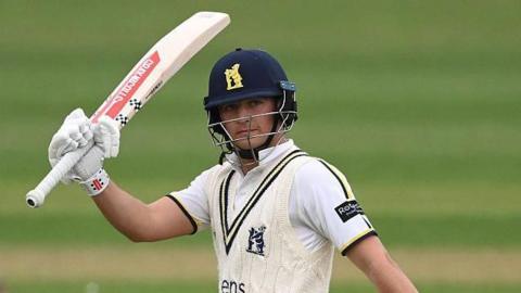Kai Smith raises his bat to the crowd after reaching his half-century for Warwickshire