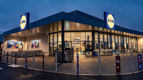A image shows the outside of a Lidl store at night time with the blue, red and yellow logo on the roof spelling out the company name. There are trolleys outside and the supermarket is lit up inside.