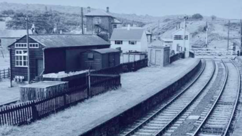 A black and white photo of a railway station. This picture has many different outbuildings surrounding the railway tracks. Next to the track is a long platform 