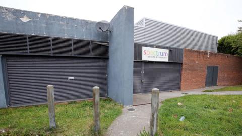 Spectrum Leisure Complex. Large black shutters have locked down the building. Grey paint is peeling from the walls.