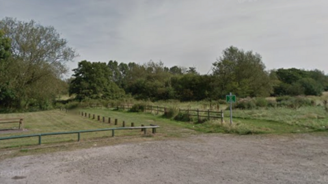 A grassy area separated by a grass path and wooden fencing. There are green trees in the background and a wooden bench on the grass to the left.
