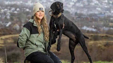 Sophie is kneeling beside Gwen and holding her up so that her two front paws are off the ground. Sophie is wearing a winter hat and coat and has long curly blonde hair. Gwen is all black with a white patch on her chest. she was long floppy ears and her shortened tail is visible. the pair are on a grassy hilltop overlooking a town 