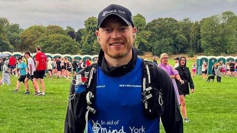 A man in front of a crowd of runners smiling and wearing a blue exercise shirt that says "York Mind" with a matching blue hat. 