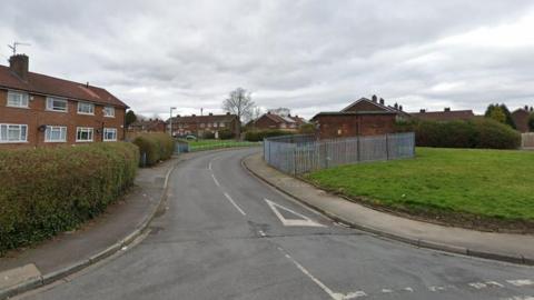 The start of Hope Hey Lane in Little Hulton, showing several two-storey red brick houses on the left and in the distance. The road bends round to right and disappears behind a small, square electricity substation surrounded by a grey, spiky metal fence.