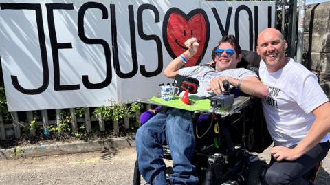 Ash Jenkins with BBC Radio Bristol breakfast presenter Joe Sims, who is wearing black jeans and a white t-shirt. Ash is wearing baggy blue jeans, a grey t-shirt, blue reflective sunglasses, and is in a motorised wheelchair. Behind them is a large white banner attached to some railings which says Jesus loves you.