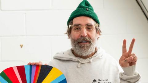 Stewart Francis Easton has a grey beard and is wearing glasses, a green wooly hat and light hoodie. He is making a two-fingered peace sign to the camera and is holding a multi-coloured piece of art he has produced. 