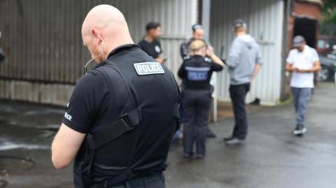 A police officer wearing a black short-sleeve shirt and vest with the word "police" stands facing away from the camera in the foreground of the picture. Other officers are in the background. They are standing in the forecourt of a car wash. The workers' identities are obscured. 