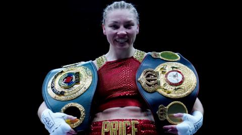 Lauren Price in her boxing kit with two large boxing champion belts, draped over each of her shoulders