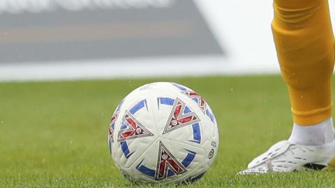 A football with National League branding in grass and a player leg standing next to it