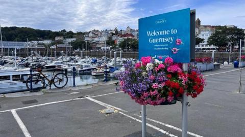 A blue Welcome to Guernsey sign with brightly-coloured flowers hanging below it, in a car park next to the island's dock.
