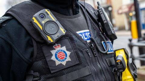 A close up of a police officer's black vest. There is a crest for Essex police on the left side and a body worn camera above it. A radio and yellow taser are also clipped to the vest.