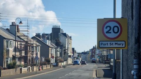 Buildings lining a tarmacked road in Port St Mary village. In the foreground there is a yellow sign with a number 20 in white circle with a red trim in the middle attached to a pole. It also reads Port St Mary in black writing on a white background.