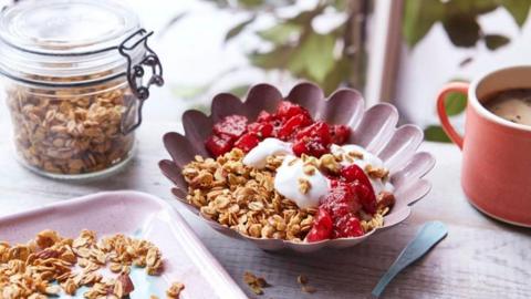 A pink bowl of granola, yoghurt and berries on a wooden surface with a jar of granola and cup of coffee