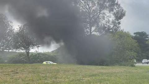 Black smoke billows from the behind the tree, which the car has crashed in to. The tree stands on a roundabout. Traffic can be seen joining the roundabout on the left. A police car has pulled over at the far side of the roundabout.