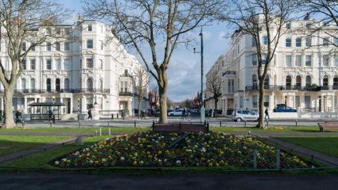 A landscaped garden with two rows of white five-storey buildings in the background. Cars and people can be seen walking and driving around the gardens which show benched and a central raised section with multi-coloured floral planting and a metalic clock hand. 