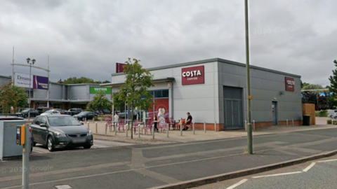 A single storey grey retail unit is photographed with larger retail units behind it. On the side of the building is a burgundy sign that reads Costa Coffee.