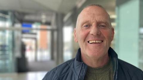 Mark Baker standing inside The Forum in Norwich. He is looking directly at the camera and smiling. He is wearing a grey jumper and dark blue coat.