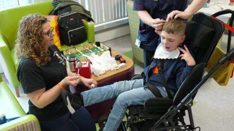 Aimee Tilley on her knees holding a spoon, wearing a black T-shirt looking at her son Isaac who is in a wheelchair in a hospital setting