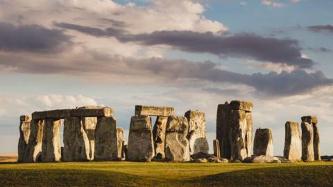 The rocks of Stonehenge stand on green grass. The sky behind them is a bright grey and there are white and dark grey cloud over them. The stones themselves are large. Some have stones lying horizontal across them 