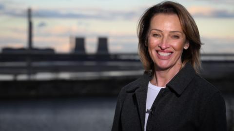 Sarah Pochin, the Reform candidate for the Runcorn and Helsby by-election standing in front of a river and some cooling towers - she has brown long hair and is wearing a white T-shirt and a black jacket