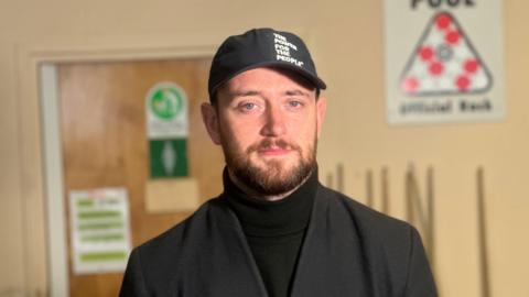 Jacob Dunne has blue eyes and a brunet beard. He is wearing a black cap that says "The Power for the People." He's wearing a black turtle neck jumper and a black blazer.