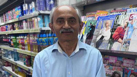 A smiling older man with a silver moustache and blue shirt is stood in the aisle of a shop with shelves full of glossy magazines and soft drinks behind him